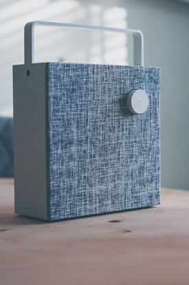 A rectangular device with a fabric-covered exterior and a circular knob is placed on a wooden surface. The device has a handle on top, suggesting portability, and it is set in a softly lit room with shadows cast on the wall.