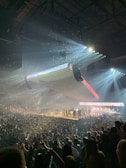 A large indoor concert with bright spotlights illuminating a stage with multiple performers. The audience is densely packed, and there is confetti in the air, creating a dynamic and festive atmosphere.