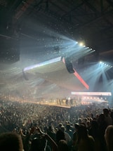 A large indoor concert with bright spotlights illuminating a stage with multiple performers. The audience is densely packed, and there is confetti in the air, creating a dynamic and festive atmosphere.
