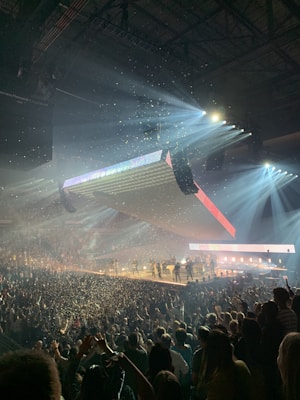 A large indoor concert with bright spotlights illuminating a stage with multiple performers. The audience is densely packed, and there is confetti in the air, creating a dynamic and festive atmosphere.
