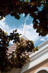 A church steeple with a cross on top is surrounded by vibrant green leaves. The steeple contrasts with a bright blue sky filled with scattered white clouds.