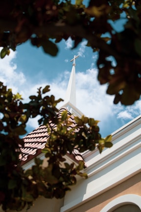 A church steeple with a cross on top is surrounded by vibrant green leaves. The steeple contrasts with a bright blue sky filled with scattered white clouds.