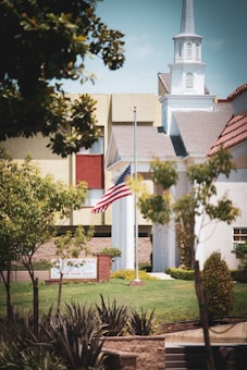 A church with a tall steeple is surrounded by trees and a well-maintained lawn. An American flag is prominently displayed on a flagpole in front of the building. The church's architecture features light-colored walls and a sloping roof.