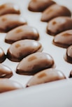 A series of glossy, oval-shaped chocolates arranged neatly in a white tray. The chocolates have a smooth, shiny surface with a rich, dark brown color.