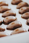 A series of glossy, oval-shaped chocolates arranged neatly in a white tray. The chocolates have a smooth, shiny surface with a rich, dark brown color.
