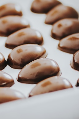 A series of glossy, oval-shaped chocolates arranged neatly in a white tray. The chocolates have a smooth, shiny surface with a rich, dark brown color.