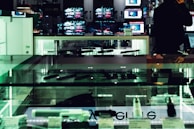 An electronics store displays multiple large screens showing images, with glass display cases containing various electronic items. The lighting is dim, casting a greenish hue over the interior. A person is visible in silhouette, seemingly engaging with a device.