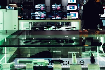 An electronics store displays multiple large screens showing images, with glass display cases containing various electronic items. The lighting is dim, casting a greenish hue over the interior. A person is visible in silhouette, seemingly engaging with a device.