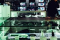 An electronics store displays multiple large screens showing images, with glass display cases containing various electronic items. The lighting is dim, casting a greenish hue over the interior. A person is visible in silhouette, seemingly engaging with a device.