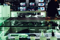 An electronics store displays multiple large screens showing images, with glass display cases containing various electronic items. The lighting is dim, casting a greenish hue over the interior. A person is visible in silhouette, seemingly engaging with a device.