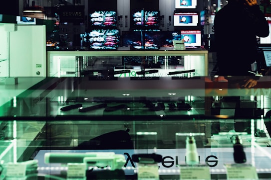 An electronics store displays multiple large screens showing images, with glass display cases containing various electronic items. The lighting is dim, casting a greenish hue over the interior. A person is visible in silhouette, seemingly engaging with a device.