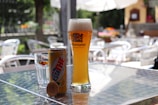 On a sunlit outdoor table, a tall glass of beer labeled 'Weihenstephaner' is placed alongside a can of 'Estathe' lemon iced tea and a glass with a 'Sanpellegrino' label. The background consists of blurred garden furniture and greenery, suggesting a casual outdoor setting.