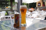 On a sunlit outdoor table, a tall glass of beer labeled 'Weihenstephaner' is placed alongside a can of 'Estathe' lemon iced tea and a glass with a 'Sanpellegrino' label. The background consists of blurred garden furniture and greenery, suggesting a casual outdoor setting.