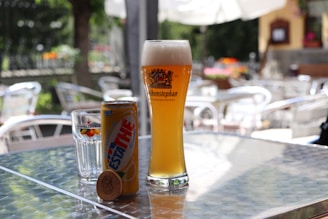 On a sunlit outdoor table, a tall glass of beer labeled 'Weihenstephaner' is placed alongside a can of 'Estathe' lemon iced tea and a glass with a 'Sanpellegrino' label. The background consists of blurred garden furniture and greenery, suggesting a casual outdoor setting.