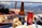 A hand is holding a fork with food from a plate filled with a meat dish and rice. In the foreground are a bottle of beer and a can of Coca-Cola on a table. The setting appears to be outdoors with a blurred background featuring a railing and mountains. A pastry or roll is partially visible on another plate.