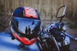 A motorcycle helmet with a red, black, and white design sits on a motorcycle. Reflections on the helmet's visor show two silhouetted figures standing nearby. The handlebar with a throttle grip and a side mirror is clearly visible. The background consists of a blurred natural setting.