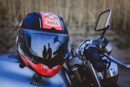 A motorcycle helmet with a red, black, and white design sits on a motorcycle. Reflections on the helmet's visor show two silhouetted figures standing nearby. The handlebar with a throttle grip and a side mirror is clearly visible. The background consists of a blurred natural setting.