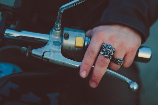 A hand is gripping a motorcycle handlebar, adorned with two detailed silver rings shaped like a skull and an intricate design. The hand appears to be wearing a dark jacket. The metal of the handlebar and the rings reflect light, giving a shiny appearance.