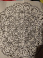 A detailed black and white mandala pattern featuring symmetrical, intricate designs with floral and geometric elements, centered on a piece of paper.