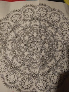 A detailed black and white mandala pattern featuring symmetrical, intricate designs with floral and geometric elements, centered on a piece of paper.
