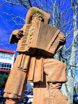 A large stone statue depicting a man wearing a cowboy hat and playing an accordion. The statue is positioned in an outdoor setting with bare trees and a building visible in the background.