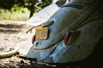 A vintage car with an old, rusted exterior is parked outdoors. The car's rear is prominently featured, showcasing a yellow license plate with the number EL 8040. The surrounding environment includes a blurred background of greenery and a tire on the ground nearby.