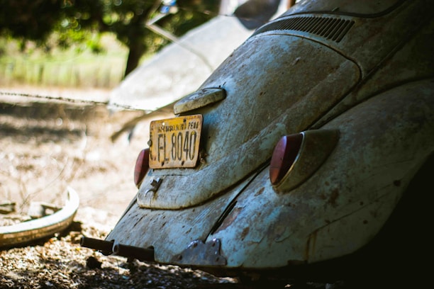 A vintage car with an old, rusted exterior is parked outdoors. The car's rear is prominently featured, showcasing a yellow license plate with the number EL 8040. The surrounding environment includes a blurred background of greenery and a tire on the ground nearby.