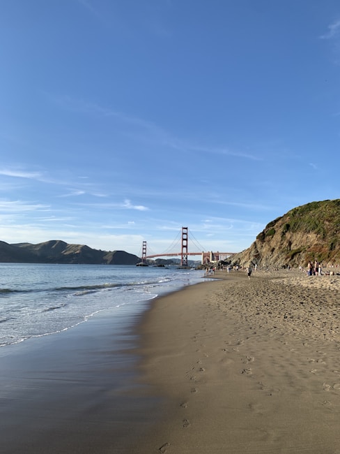 A serene beach scene with gentle waves lapping against the shore. In the background, the iconic Golden Gate Bridge stretches across the water under a clear blue sky. People stroll along the sandy beach, leaving footprints behind, while others relax near the rocky cliffs that line the edge.