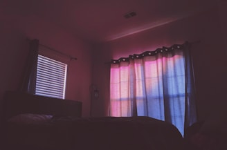 A dimly lit room features a bed with a partially visible pillow and blanket. The window is covered with curtains displaying a gradient of soft pink and blue hues, likely from the sunset, casting a calming glow across the room. A closed blinds window on the left side of the room allows a faint light to filter through its slats. A lantern-style wall sconce is visible between the windows.