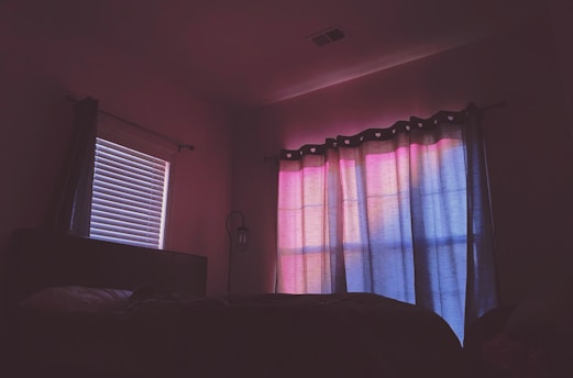 A dimly lit room features a bed with a partially visible pillow and blanket. The window is covered with curtains displaying a gradient of soft pink and blue hues, likely from the sunset, casting a calming glow across the room. A closed blinds window on the left side of the room allows a faint light to filter through its slats. A lantern-style wall sconce is visible between the windows.