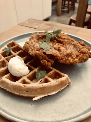 A plate featuring a waffle topped with crispy fried chicken garnished with cilantro leaves and a dollop of creamy sauce, set on a wooden table in a cozy dining environment.