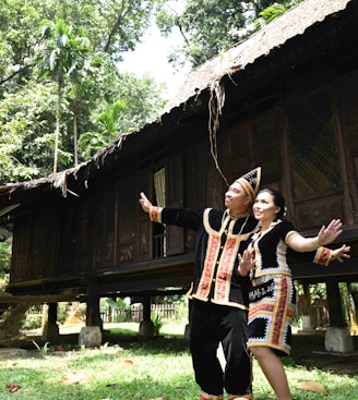 man and woman in black-and-brown matching robes standing side by side in front of wooden house