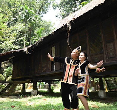 man and woman in black-and-brown matching robes standing side by side in front of wooden house