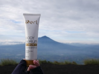 A hand wearing gloves holds a tube of body serum labeled 'erl Body Serum Wow Lightening' with a distant mountainous landscape in the background under a cloudy sky.