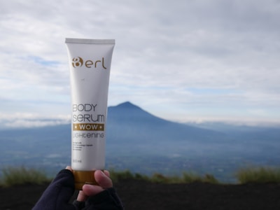 A hand wearing gloves holds a tube of body serum labeled 'erl Body Serum Wow Lightening' with a distant mountainous landscape in the background under a cloudy sky.