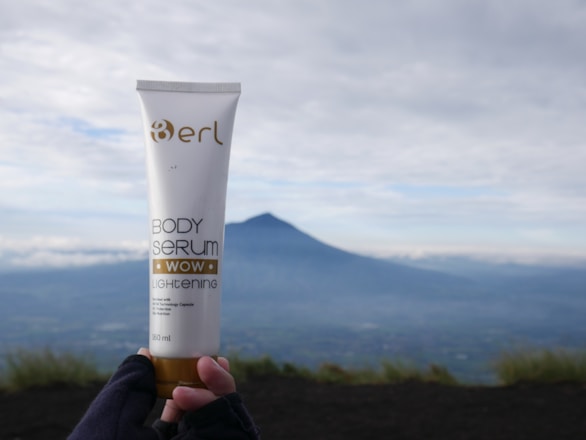 A hand wearing gloves holds a tube of body serum labeled 'erl Body Serum Wow Lightening' with a distant mountainous landscape in the background under a cloudy sky.