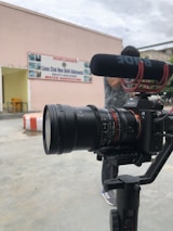 A professional camera setup is positioned in the foreground, equipped with a microphone. In the background, there's a building with a sign that reads 'Lions Club New Delhi Alaknanda Water Harvesting'. A person is partially visible behind the camera.