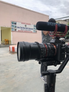 A professional camera setup is positioned in the foreground, equipped with a microphone. In the background, there's a building with a sign that reads 'Lions Club New Delhi Alaknanda Water Harvesting'. A person is partially visible behind the camera.