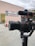 A professional camera setup is positioned in the foreground, equipped with a microphone. In the background, there's a building with a sign that reads 'Lions Club New Delhi Alaknanda Water Harvesting'. A person is partially visible behind the camera.