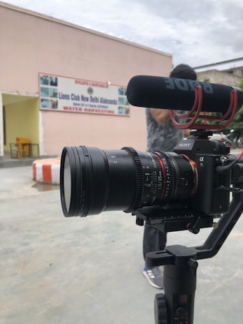 A professional camera setup is positioned in the foreground, equipped with a microphone. In the background, there's a building with a sign that reads 'Lions Club New Delhi Alaknanda Water Harvesting'. A person is partially visible behind the camera.