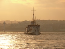 A ship sails on calm waters under a golden sunset. The background features a silhouetted city skyline with a pale sky and faint outlines of buildings.