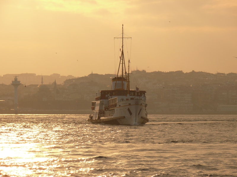 A ship sails on calm waters under a golden sunset. The background features a silhouetted city skyline with a pale sky and faint outlines of buildings.