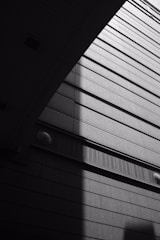 A black and white architectural photograph featuring the geometric lines and angles of a building. The image captures strong contrasts between light and shadow, highlighting the texture of the exterior surfaces and the pattern of lines.