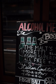 A chalkboard menu showcases various drink options, with hand-drawn illustrations of beer bottles and decorative text. It lists beverages such as 'Moretti' and 'Asahi Super Dry'. The menu features a vintage, artisanal design, with flourishes and cursive text in both English and Japanese.