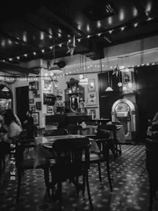 A dimly lit interior of a vintage-style cafe or bar, featuring wooden tables and chairs, a classic jukebox, and decorative elements such as hanging star-shaped lights and framed pictures on the walls. The floor has a patterned design, and strings of lights hang from the ceiling creating a cozy and nostalgic atmosphere.