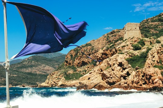A large, blue flag billows dramatically in the wind, mounted on a pole near the edge of a rugged coastline. The background features rocky cliffs with patches of green vegetation and an old stone tower perched on a hill. Waves crash against the shore, and the clear sky is a bright shade of blue.