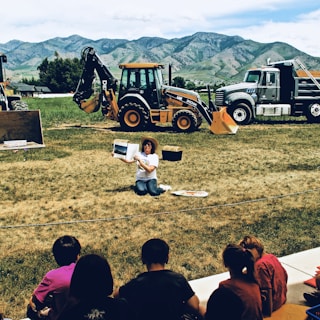 A person sitting on the grass, holding up a book and apparently reading to a group of children seated in front of them. Behind the person, there are construction vehicles, including a backhoe and a dump truck. The background consists of green grassy fields and mountains.