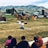 A person sitting on the grass, holding up a book and apparently reading to a group of children seated in front of them. Behind the person, there are construction vehicles, including a backhoe and a dump truck. The background consists of green grassy fields and mountains.