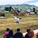 A person sitting on the grass, holding up a book and apparently reading to a group of children seated in front of them. Behind the person, there are construction vehicles, including a backhoe and a dump truck. The background consists of green grassy fields and mountains.