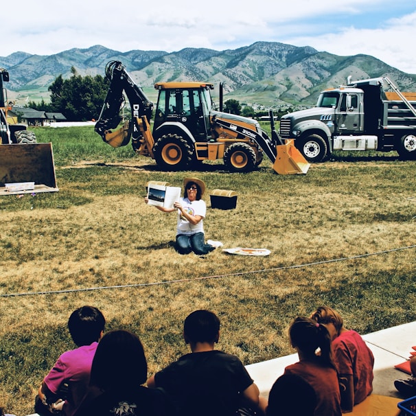 A group of kids gathered around a volunteer reading a storybook under a large tree